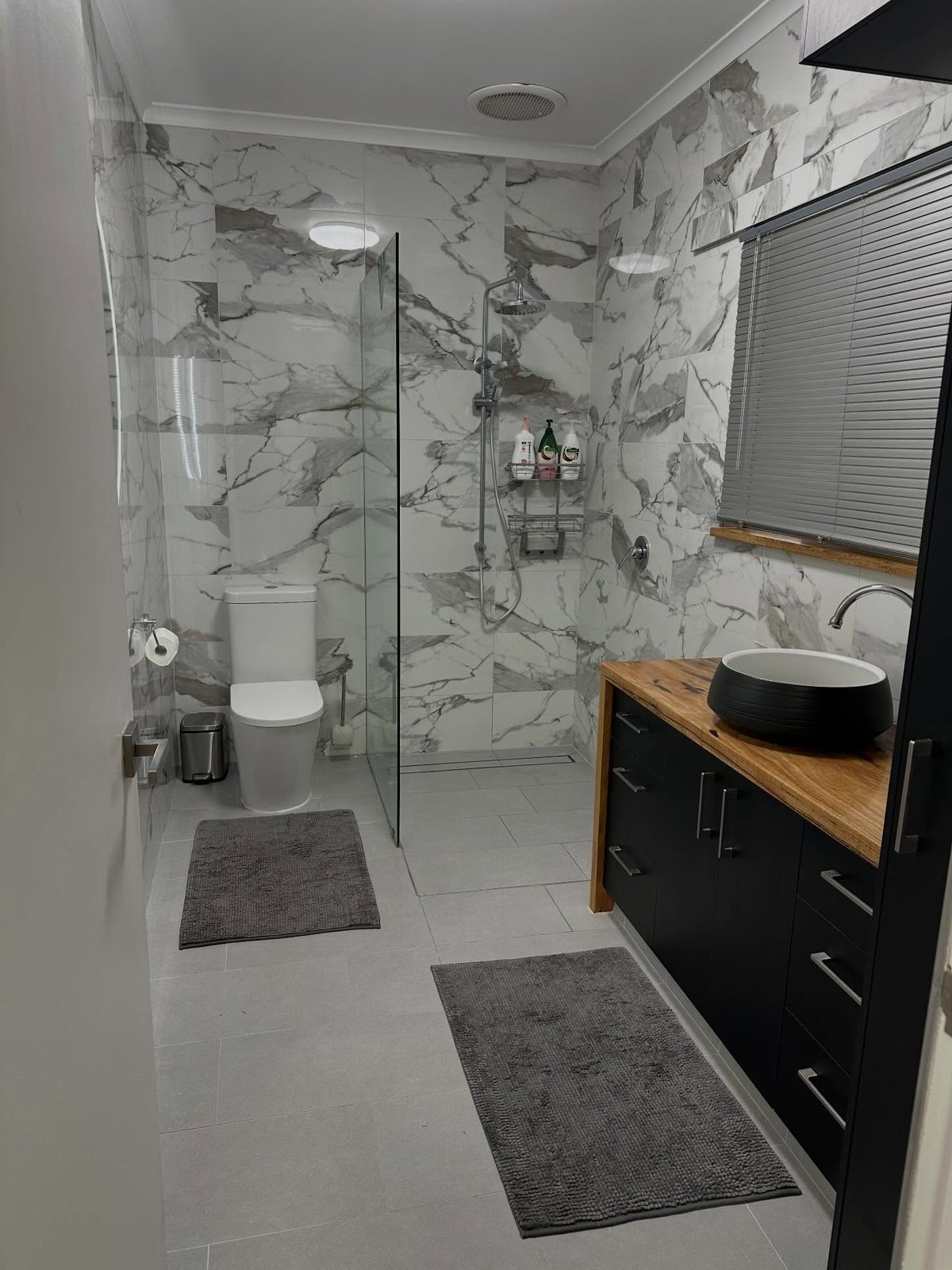 After: the same bathroom with marble-look walls, a black vanity with a timber top and a walk-in shower